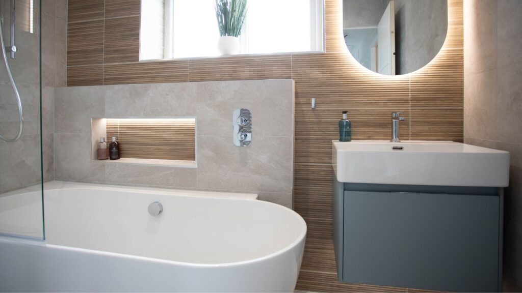 A modern bathroom with a white oval bathtub and a light blue floating vanity. The room has light-toned textured tile and a round backlit mirror.