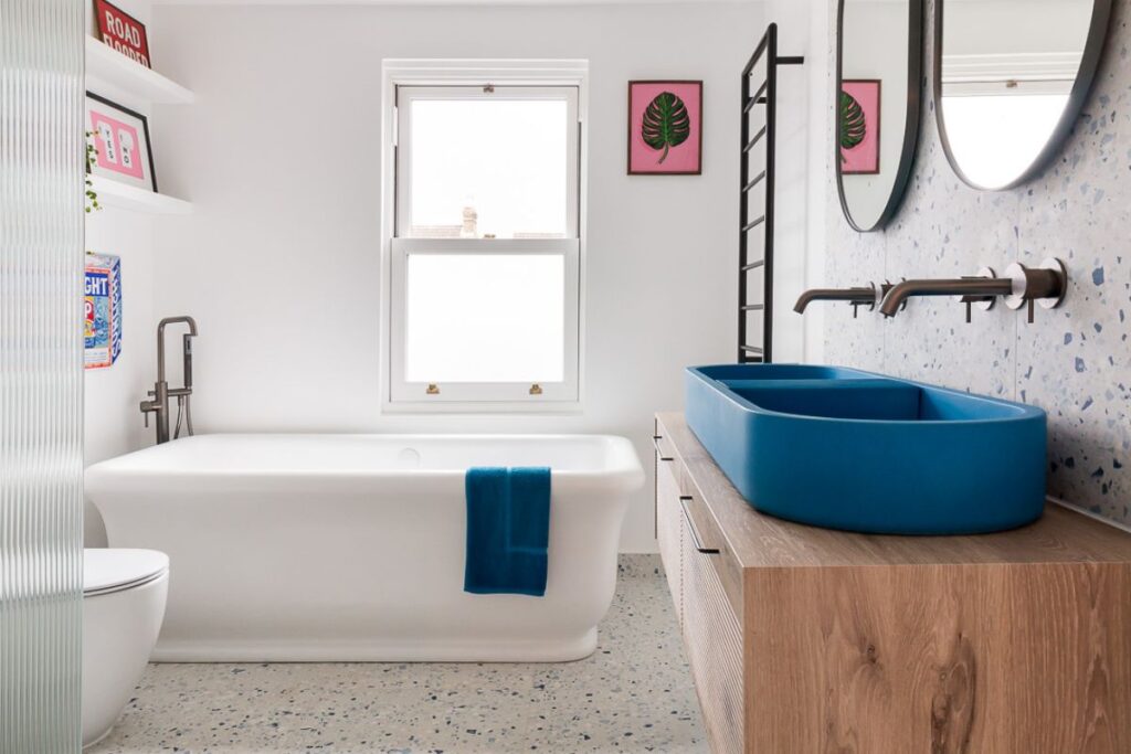 A modern bathroom featuring a vibrant blue double sink on a wooden vanity, a white bathtub with a matching blue towel, and blue-flecked terrazzo flooring.