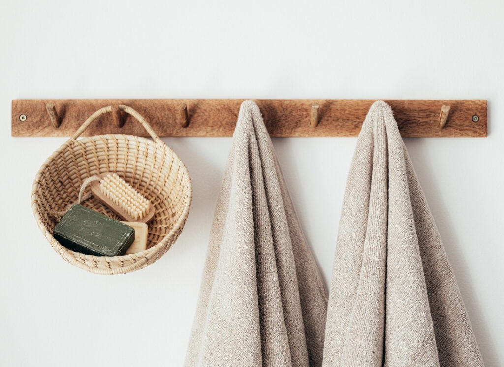 A wooden peg rail on a white wall holding a small wicker basket with soap and a brush, alongside two neutral-toned towels.